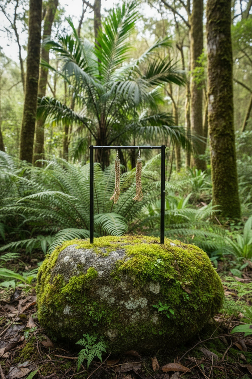 A pair of long braided tassel earrings in brown and beige hanging from a wooden display stand against a bright green background.
