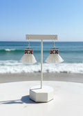 A pair of white tassel earrings with a red, black, and white patterned band, displayed on a white stand set against a beach background with the ocean and blue sky.
