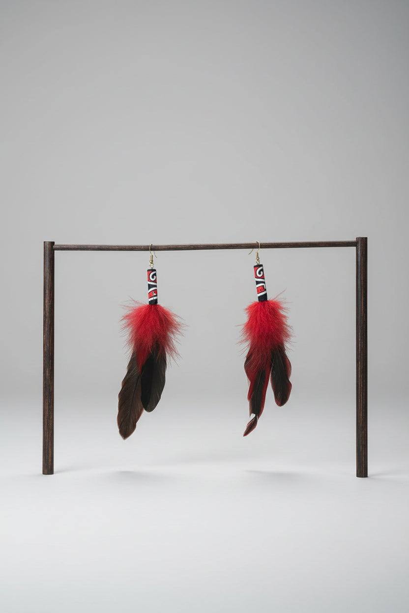 A pair of handmade dangle earrings with bright red feathers and black feathers, hanging from a white PVC pipe frame against a green fabric backdrop.