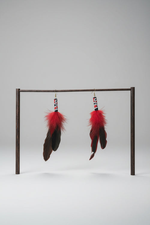 A pair of handmade dangle earrings with bright red feathers and black feathers, hanging from a white PVC pipe frame against a green fabric backdrop.
